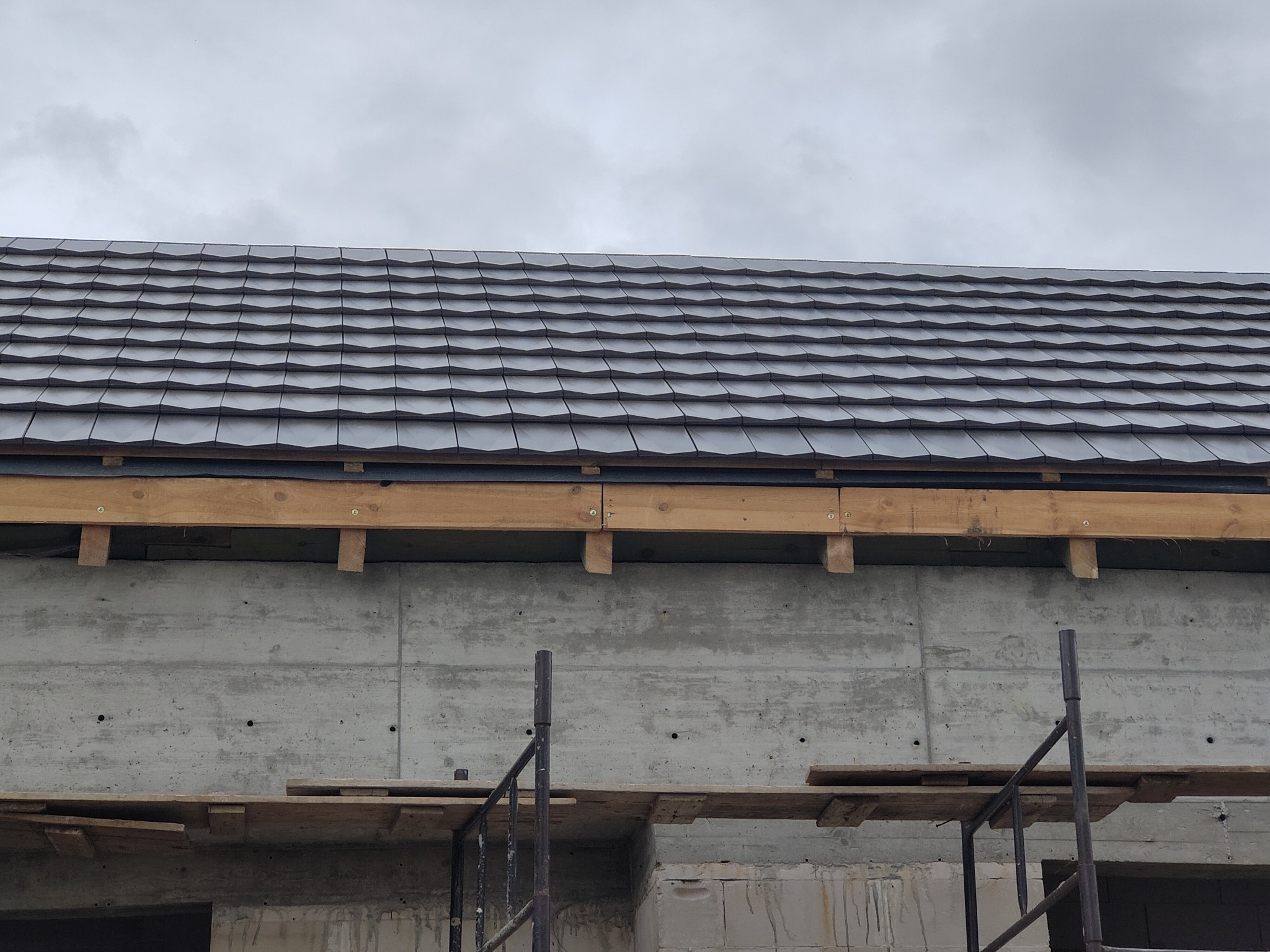 Szare płytki dachowe ułożone na nowym budynku z betonu, widoczne rusztowania i drewniane elementy konstrukcyjne.
