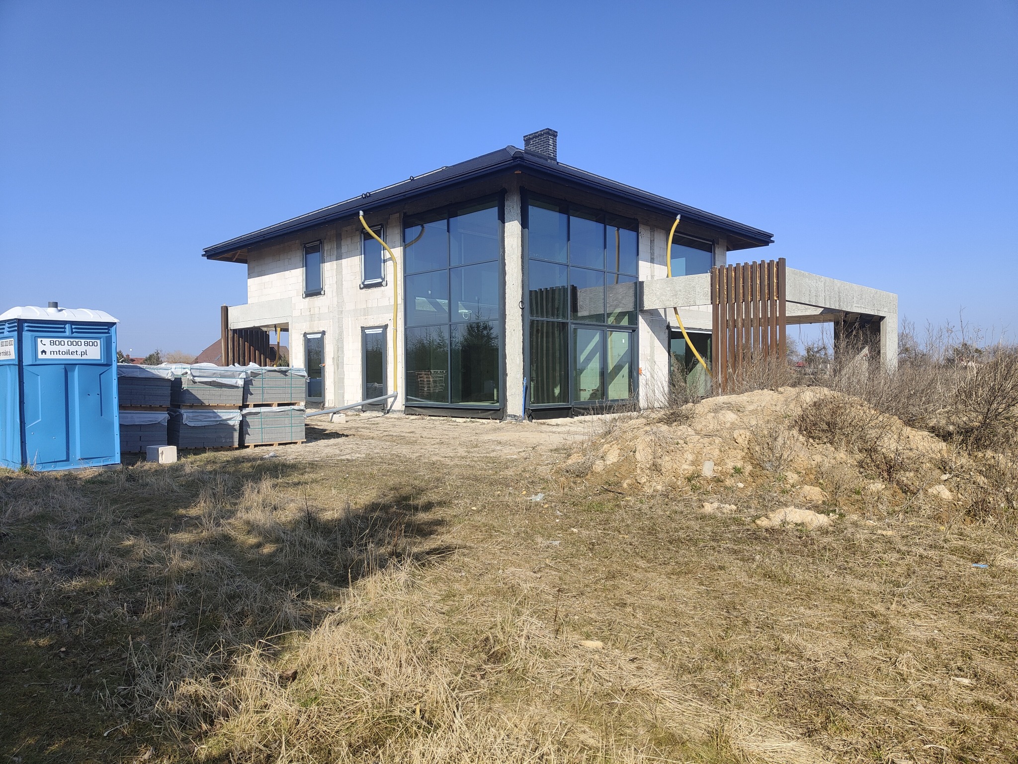 Budowa nowoczesnego domu jednorodzinnego z dużymi przeszkleniami, widoczne pustaki, elementy konstrukcyjne balkonu, prowizoryczna toaleta i materiały budowlane na działce.