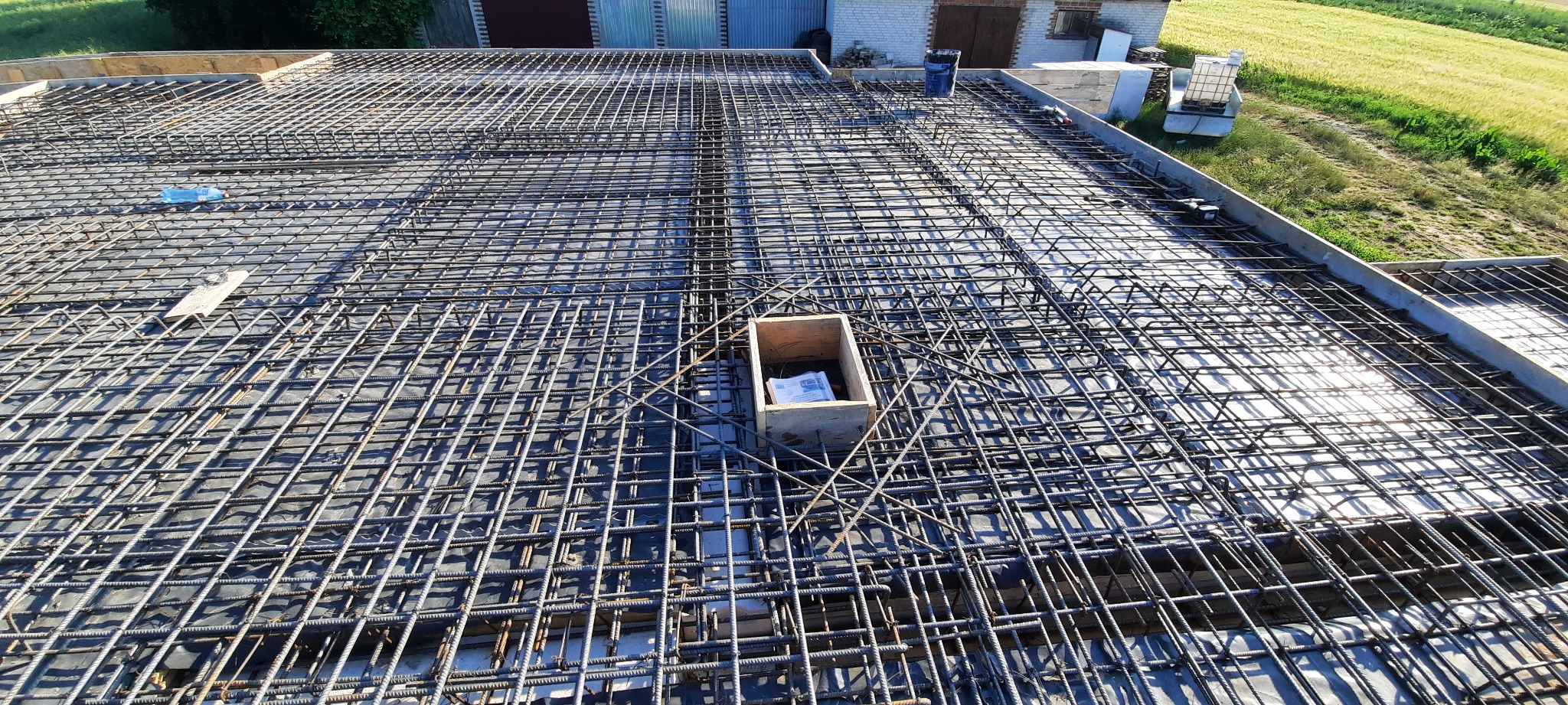 Zbrojenie pod wylewkę na przygotowanym szalunku, widoczna siatka z prętów stalowych i drewniana skrzynka z dokumentami w rogu. W tle budynek gospodarczy i pole.