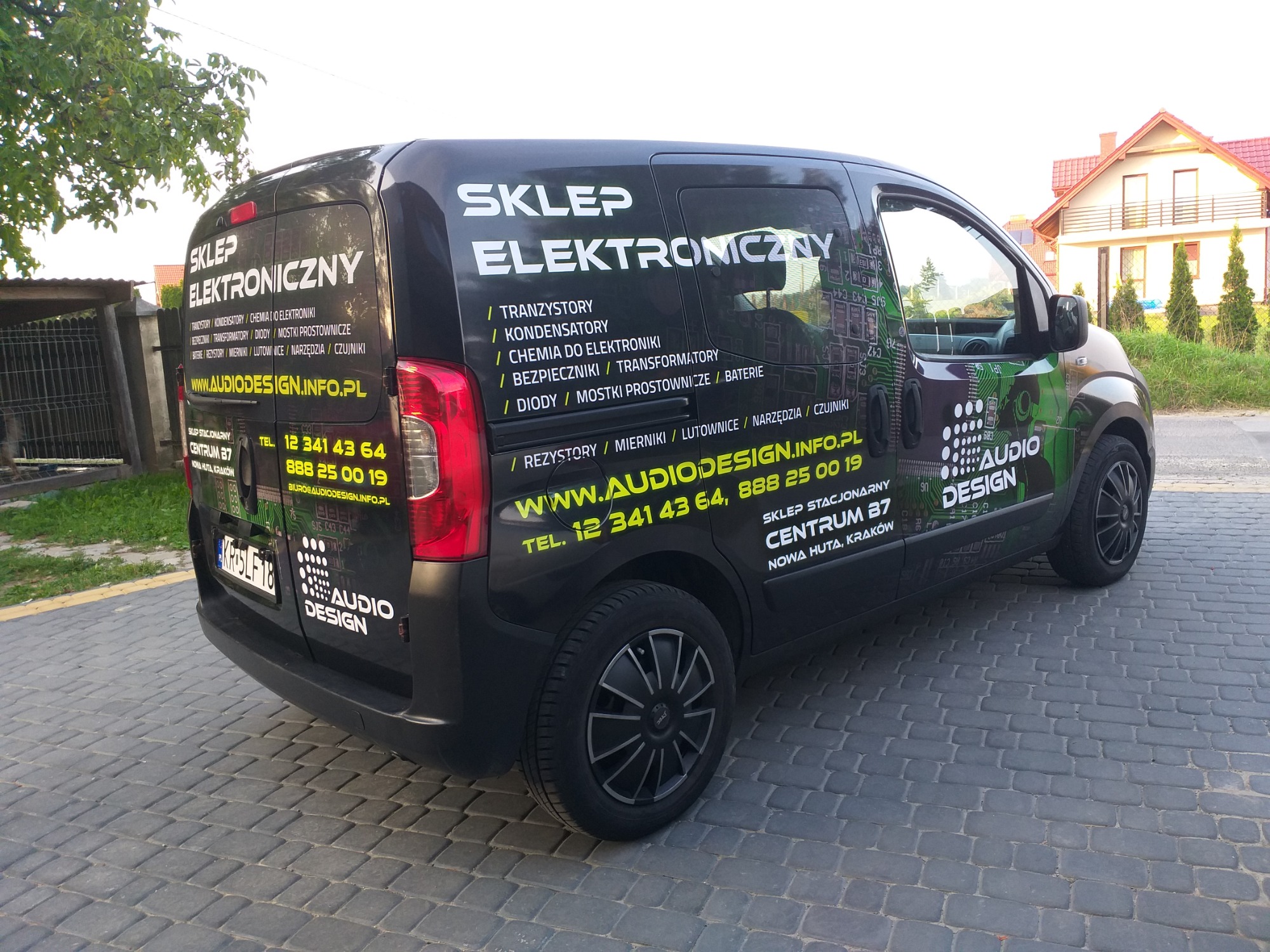 Czarny samochód dostawczy z reklamą sklepu elektronicznego Audio Design, prezentujący listę komponentów elektronicznych i dane kontaktowe, zaparkowany na brukowanej ulicy.