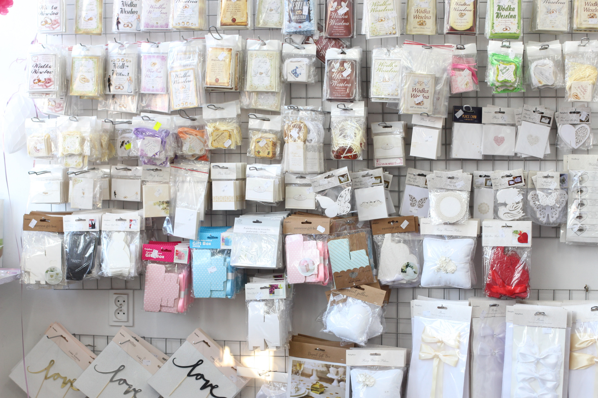 Ściana z ekspozycją artykułów weselnych w folii: zaproszenia, zawieszki na alkohol, pudełeczka na upominki, poduszki na obrączki, dekoracje tortów z napisem 'love'.