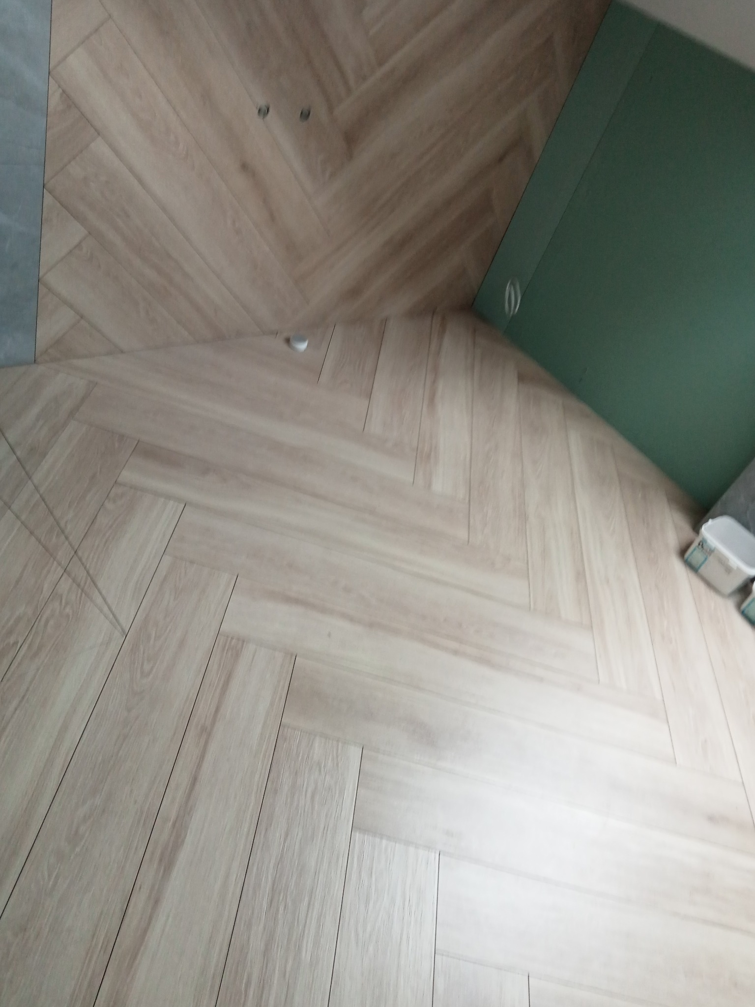 Ukończona podłoga z paneli ułożonych w jodełkę francuską, z fragmentem zielonej ściany z płyty karton gips w rogu, widoczne puszki instalacyjne w suficie i wiadro po farbie przy ścianie.