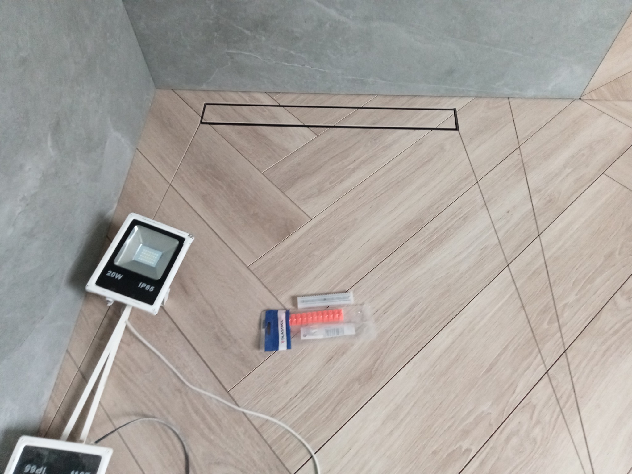 Ukończona podłoga w łazience z jasnych płytek ułożonych w jodełkę, z czarnym odpływem liniowym, oświetlona lampą roboczą i z widocznymi akcesoriami do poziomowania płytek.