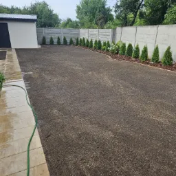 Świeżo zasiana trawa na posesji z betonowym ogrodzeniem, obok budynku gospodarczego i chodnika, z widocznym wężem ogrodowym