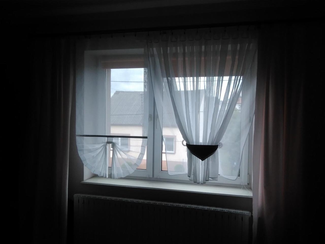 Okno z białymi, asymetrycznie upiętymi zasłonami, widok na budynek z szarą dachówką
