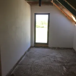 Pokój na poddaszu po nałożeniu gładzi na ściany, widoczne instalacje pod sufitem i okno z widokiem na zieleń.