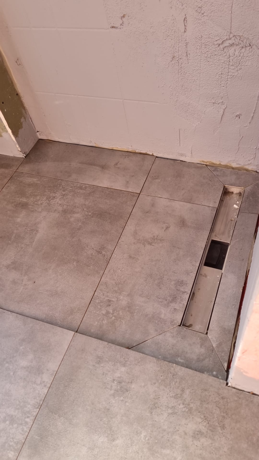 Ułożona szara, matowa podłoga z odpływem liniowym w łazience. Widoczne niedokończone ściany z gładzią. Minimalistyczny design.