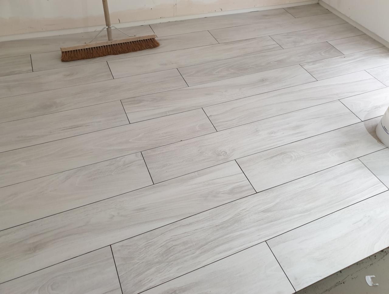 Ułożona podłoga z jasnych, drewnopodobnych płytek ceramicznych z ciemną fugą, widoczna szczotka do zamiatania i wiadro z farbą w rogu pomieszczenia.