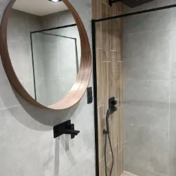 Nowoczesna łazienka z okrągłym lustrem w drewnianej ramie, czarną armaturą i kabiną prysznicową z deszczownicą, wykończona płytkami w odcieniach szarości i drewna.