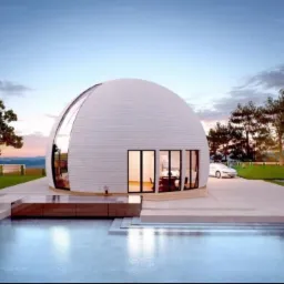 Nowoczesny dom kopułowy z białymi panelami, dużym panoramicznym oknem i basenem, otoczony zielenią i drzewami, z samochodem zaparkowanym na podjeździe.