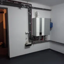 Nowoczesny, szaro-biały wiszący piec z podłączonymi rurami w schludnej kotłowni z szarym podłożem i widoczną instalacją rur pod sufitem.