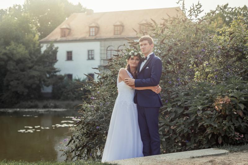 Para młoda w objęciach na tle jeziora i zabytkowego budynku z czerwonym dachem. Panna młoda w wianku, pan młody w granatowym garniturze.