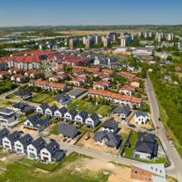 Zdjęcie lotnicze osiedla domów jednorodzinnych w Pławach, z widocznymi nowymi i starszymi budynkami, oraz zielenią i zabudową wielorodzinną w tle.