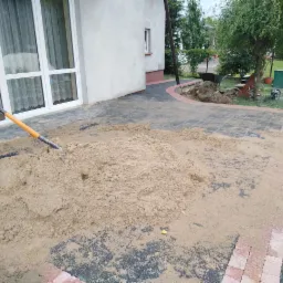 Stos piasku z wbita łopatą na częściowo ułożonej nawierzchni z kostki brukowej w kolorach szarości i czerwieni, widoczne prace ziemne i przygotowanie podłoża.
