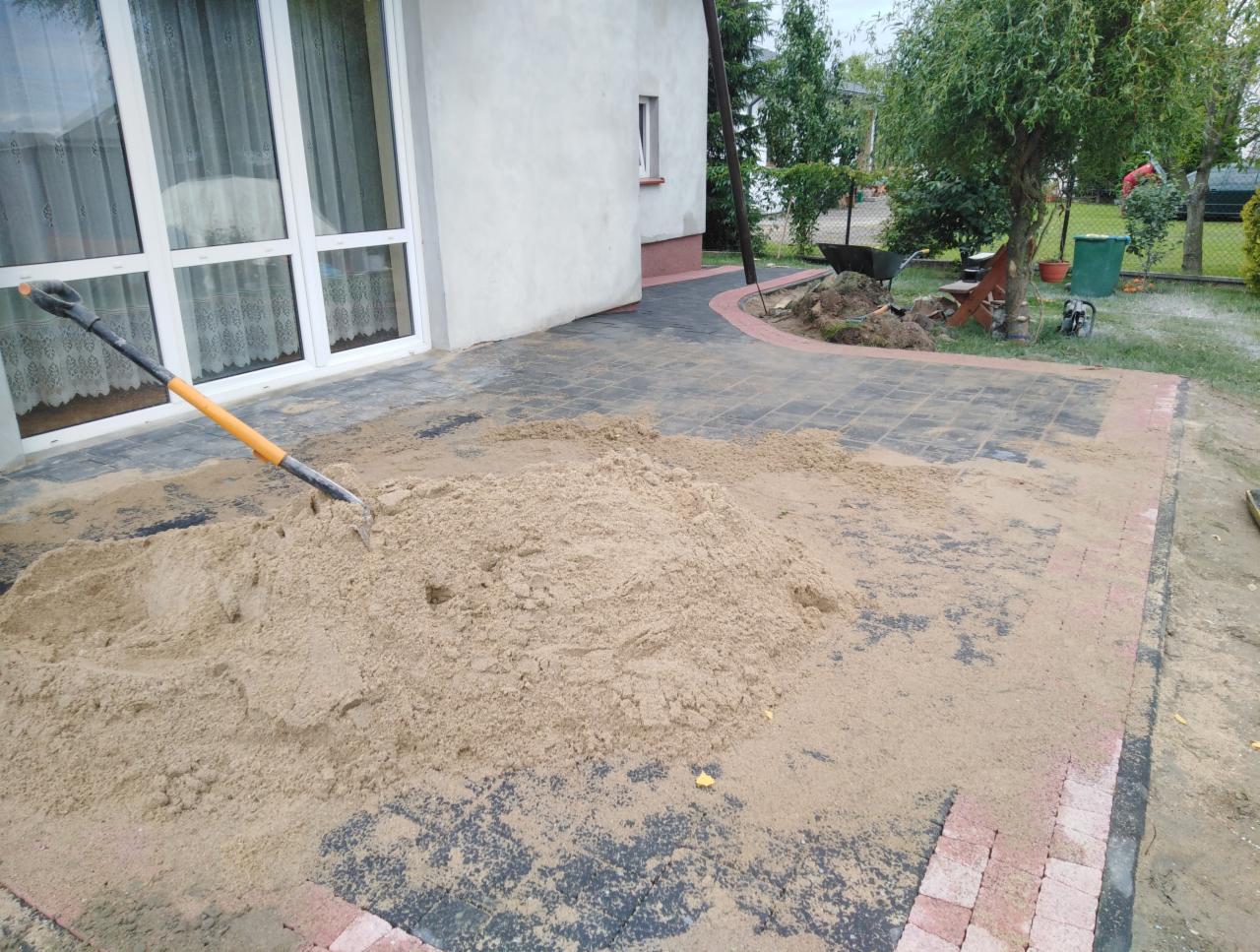 Stos piasku z wbita łopatą na częściowo ułożonej nawierzchni z kostki brukowej w kolorach szarości i czerwieni, widoczne prace ziemne i przygotowanie podłoża.
