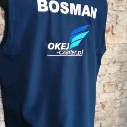 Granatowa kamizelka z białym napisem 'BOSMAN' i logo firmy 'OKEJ-czarter.pl', widoczna z tyłu, na tle ceglanej ściany.