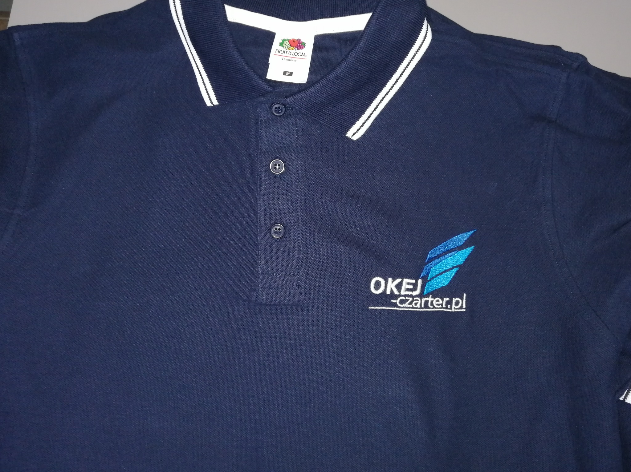 Granatowa koszulka polo z białym kołnierzykiem, haftowanym logo firmy 'OKEJ -czarter.pl' w kolorze niebieskim, widoczne logo producenta 'Fruit of the Loom'.
