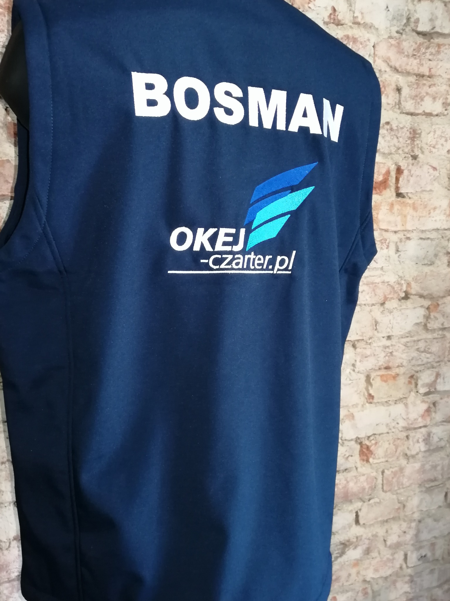 Granatowa kamizelka z białym napisem 'BOSMAN' i logo firmy 'OKEJ-czarter.pl', widoczna z tyłu, na tle ceglanej ściany.
