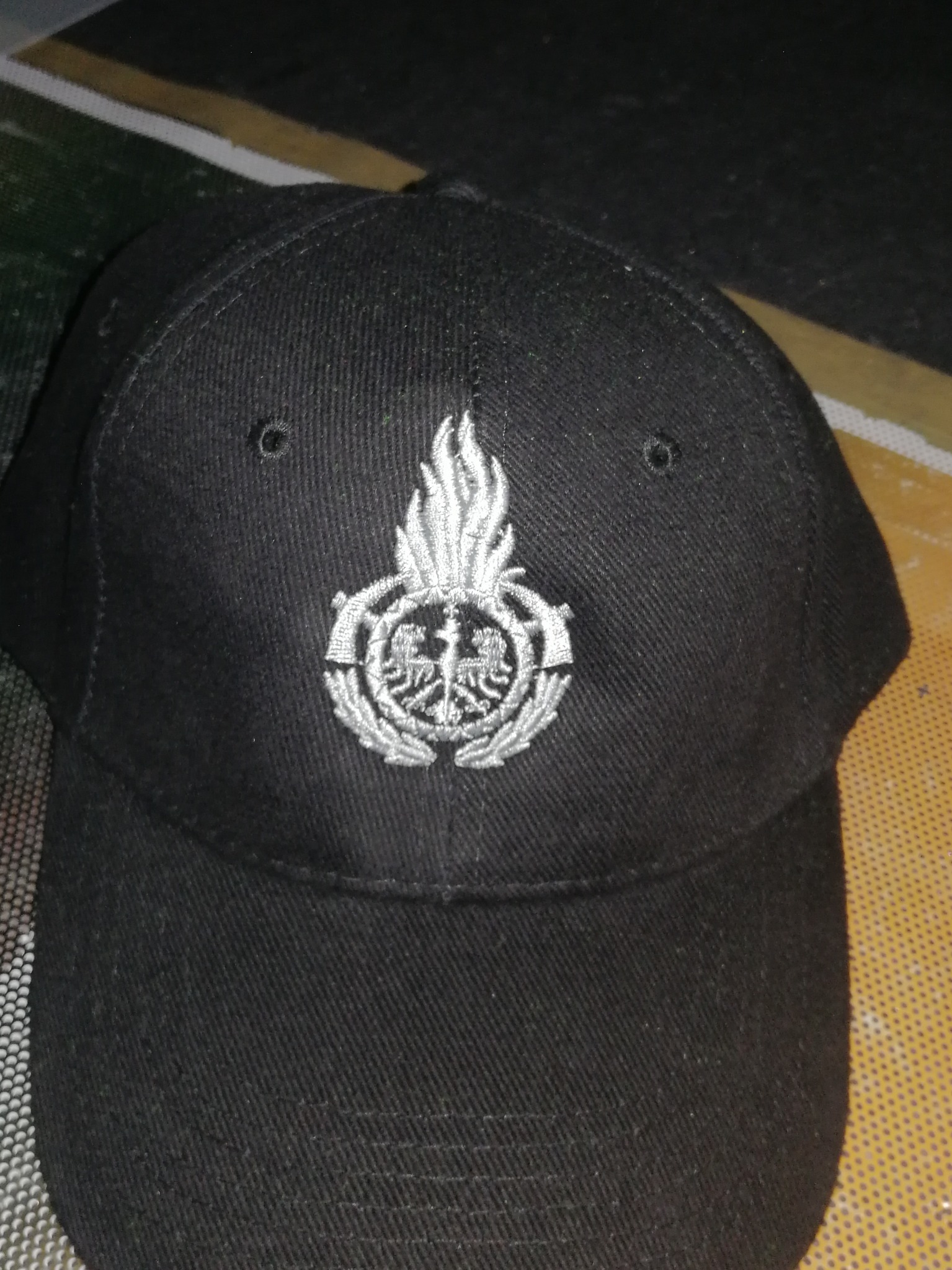 Czarna czapka bejsbolówka z wyhaftowanym srebrnym orłem w koronie otoczonym wieńcem i płomieniem na froncie.