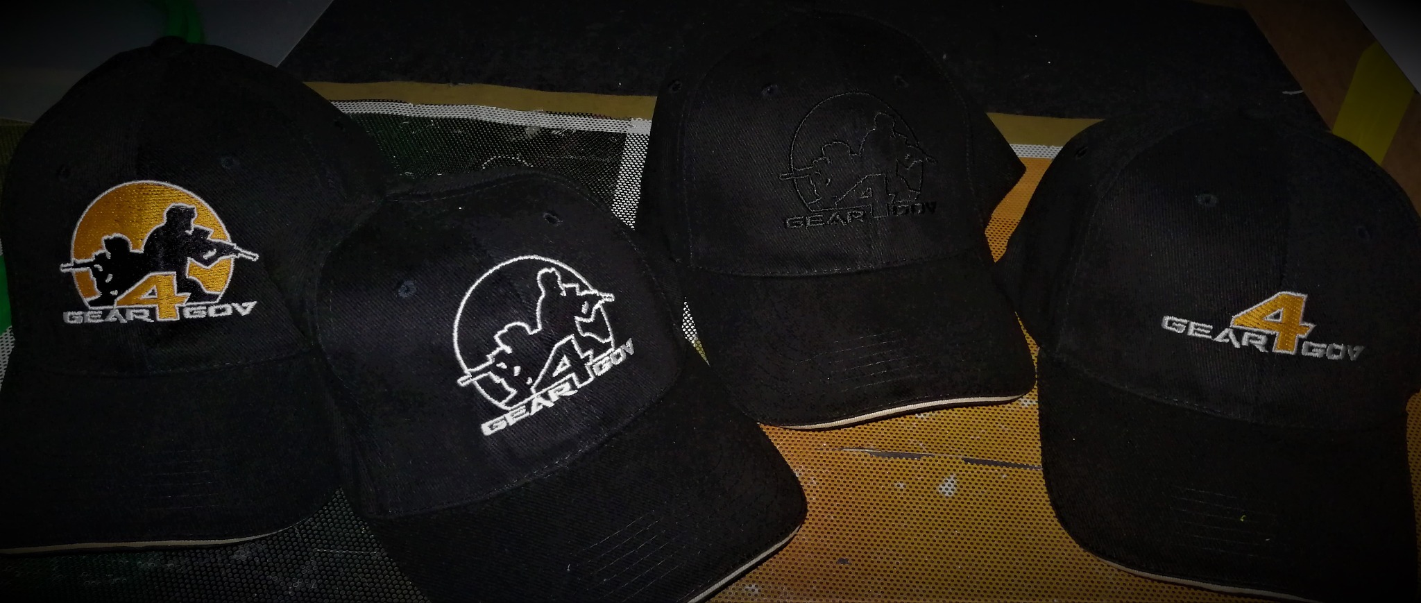 Cztery czarne czapki z daszkiem z różnymi wariantami haftowanego logo 'GEAR4GOV' z sylwetkami żołnierzy, leżące na perforowanej powierzchni.
