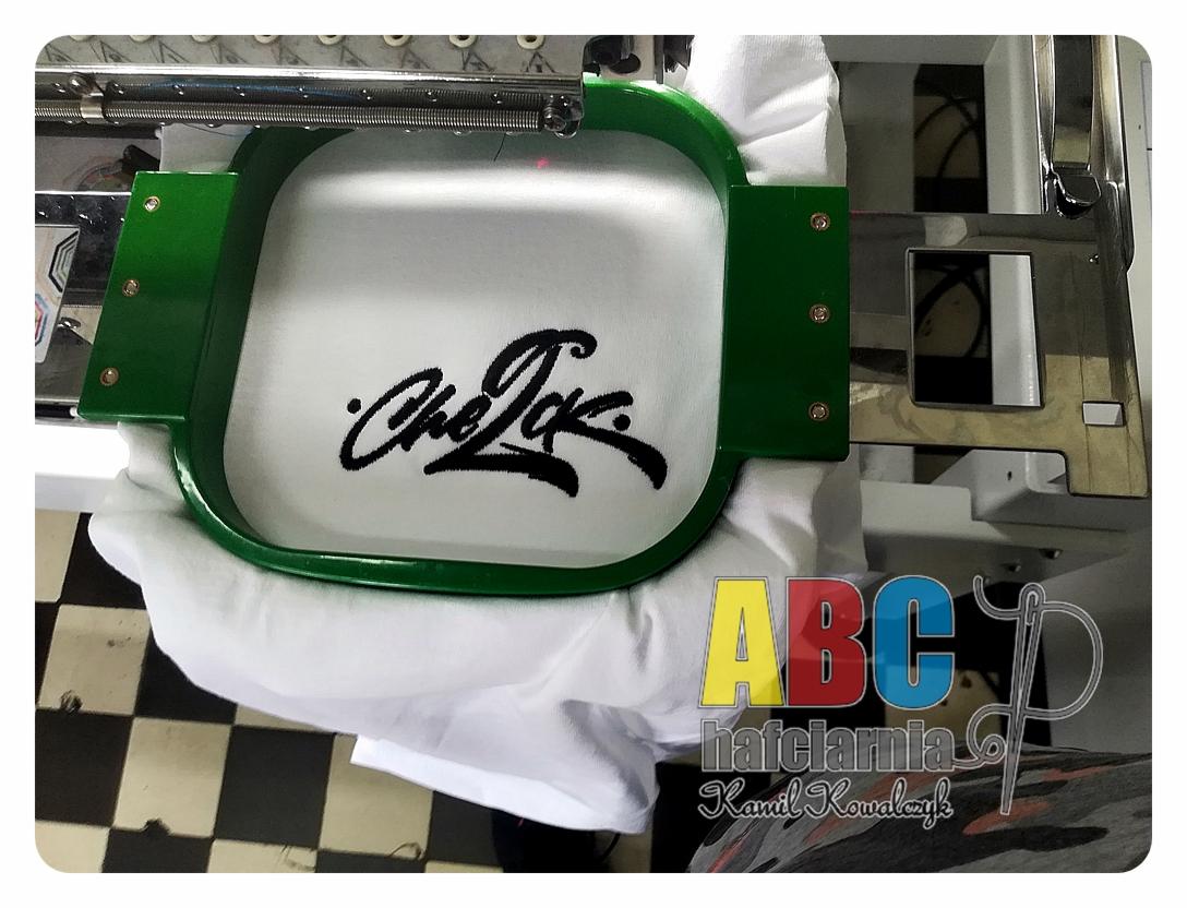 Haftowany czarny napis 'Che Jak' na białej tkaninie, umieszczony w zielonym ramie hafciarki, widok z bliska na etapie tworzenia.