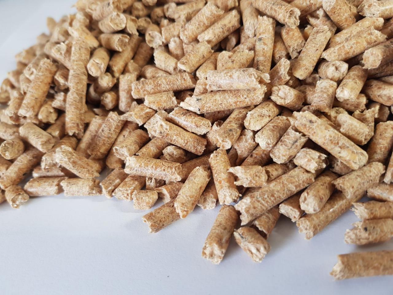 Stos brązowych, cylindrycznych brykietów z biomasy na jasnej powierzchni, widoczne drobne fragmenty i tekstura drewna.