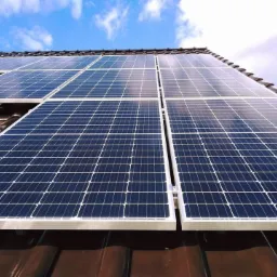 Panele fotowoltaiczne zamontowane na brązowym dachu z dachówki ceramicznej, widok pod kątem w słoneczny dzień z błękitnym niebem i białymi chmurami.