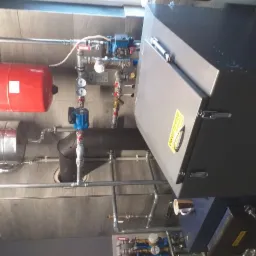 Kompletna instalacja grzewcza z piecem, zbiornikiem wyrównawczym (czerwony) i zasobnikiem ciepłej wody (srebrny), widoczne liczne rury i zawory, zamontowana na ścianie wyłożonej płytkami.