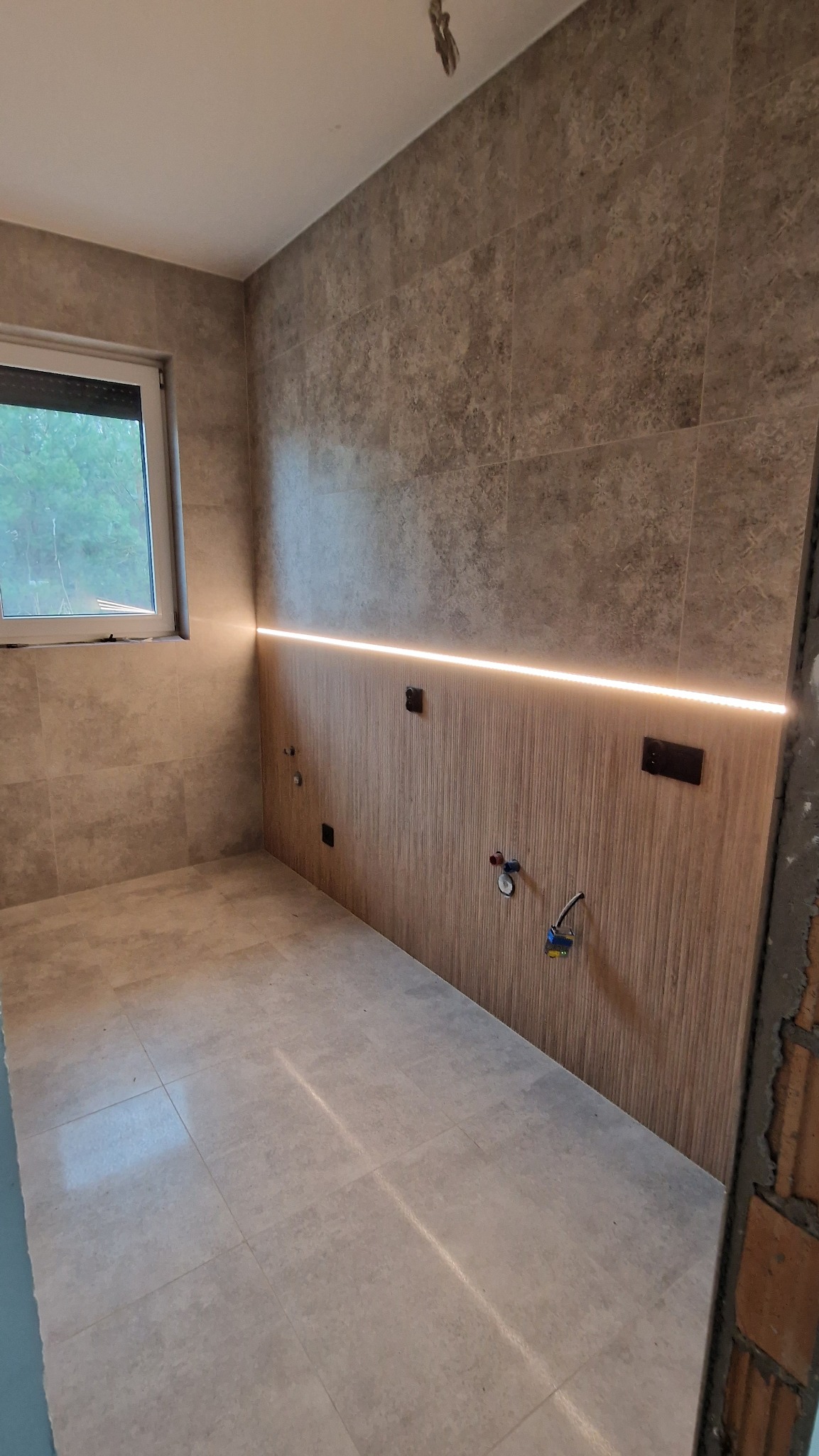 Łazienka w trakcie remontu z położonymi szarymi płytkami podłogowymi i częściowo wykończonymi ścianami z podświetleniem LED oraz wystającymi przewodami instalacji wodnej i elektrycznej.