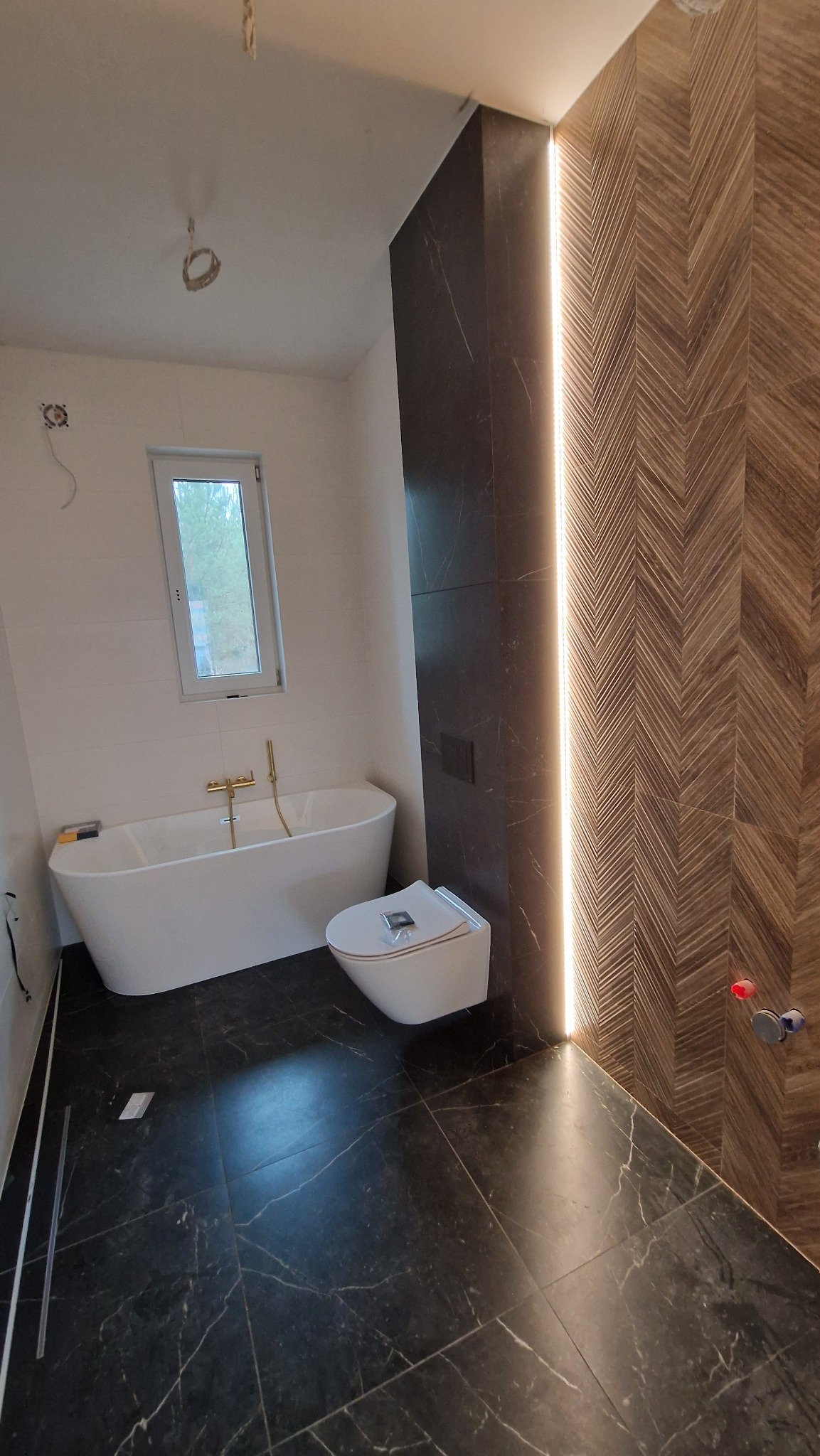 Nowoczesna łazienka w trakcie wykańczania: czarna marmurowa podłoga, biała wanna wolnostojąca z złotą armaturą, wisząca toaleta, ściana z drewnopodobnych płytek ułożonych w jodełkę z podświetleniem...