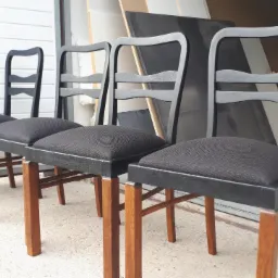 Cztery odnowione krzesła z ciemnoszarymi siedziskami i drewnianymi nogami, ustawione w rzędzie, z widocznym tłem warsztatowym i narzędziami.