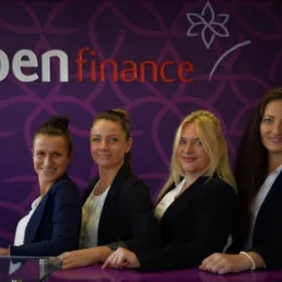 Cztery uśmiechnięte kobiety w biurze Open Finance, stojące za purpurowym biurkiem z logo firmy na ścianie w tle.