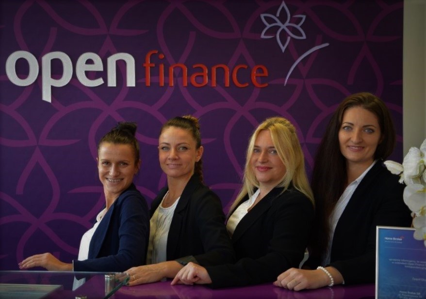 Cztery uśmiechnięte kobiety w biurze Open Finance, stojące za purpurowym biurkiem z logo firmy na ścianie w tle.