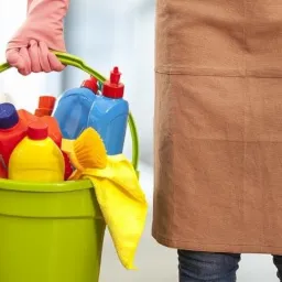 Osoba w brązowym fartuchu trzyma zielone wiadro wypełnione środkami czystości, w tym butelkami, spryskiwaczem i żółtą ściereczką; różowe rękawice ochronne na dłoni