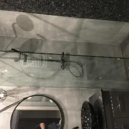 Nowoczesna łazienka z szarymi płytkami, czarną armaturą, szklaną kabiną prysznicową i okrągłym lustrem z widocznym odbiciem osoby.