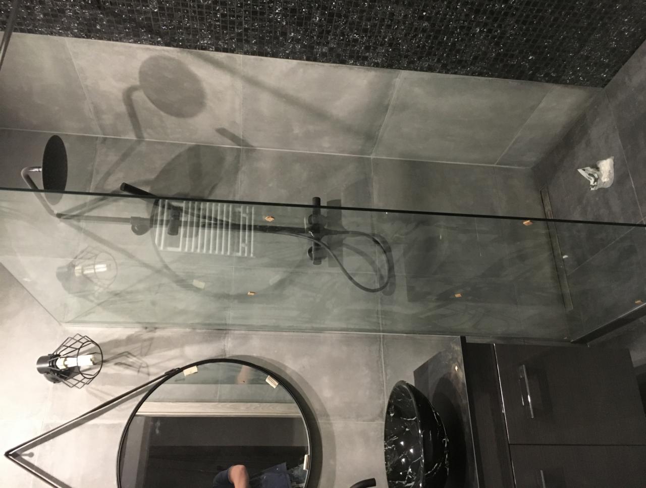 Nowoczesna łazienka z szarymi płytkami, czarną armaturą, szklaną kabiną prysznicową i okrągłym lustrem z widocznym odbiciem osoby.