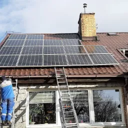 Montaż paneli słonecznych na dachu domu z ceglaną elewacją; pracownik na drabinie przy rynnie, widoczne okna z odbiciami drzew.