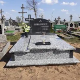 Granitowy pomnik z ciemnoszarym nagrobkiem, kwiatami w czarnym wazonie i zniczem, na tle cmentarza z krzyżami i turbiną wiatrową w oddali.