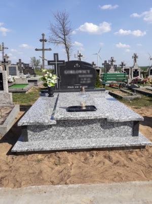 Granitowy pomnik z ciemnoszarym nagrobkiem, kwiatami w czarnym wazonie i zniczem, na tle cmentarza z krzyżami i turbiną wiatrową w oddali.