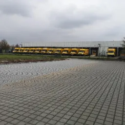 Szeroki plac wyłożony szarą kostką brukową typu ażur, przed budynkiem magazynowym z zaparkowanymi żółtymi samochodami dostawczymi.