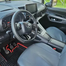 Wnętrze samochodu Toyota Proace City z szarymi pokrowcami na siedzeniach i czarno-czerwonymi dywanikami, widok z perspektywy kierowcy.