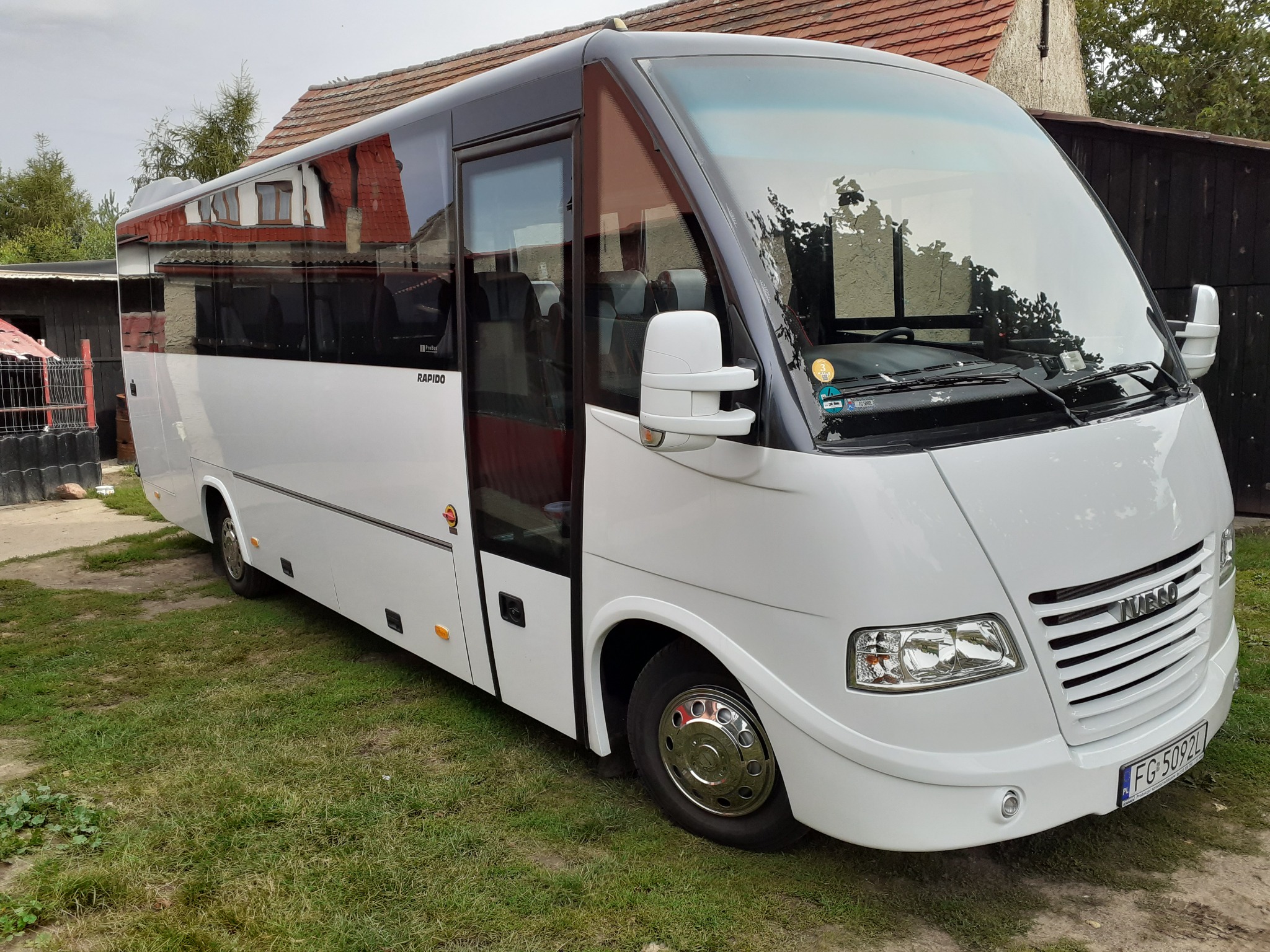 Biały minibus marki Iveco Rapido z otwartymi drzwiami, zaparkowany na trawie przed budynkiem z czerwoną dachówką.