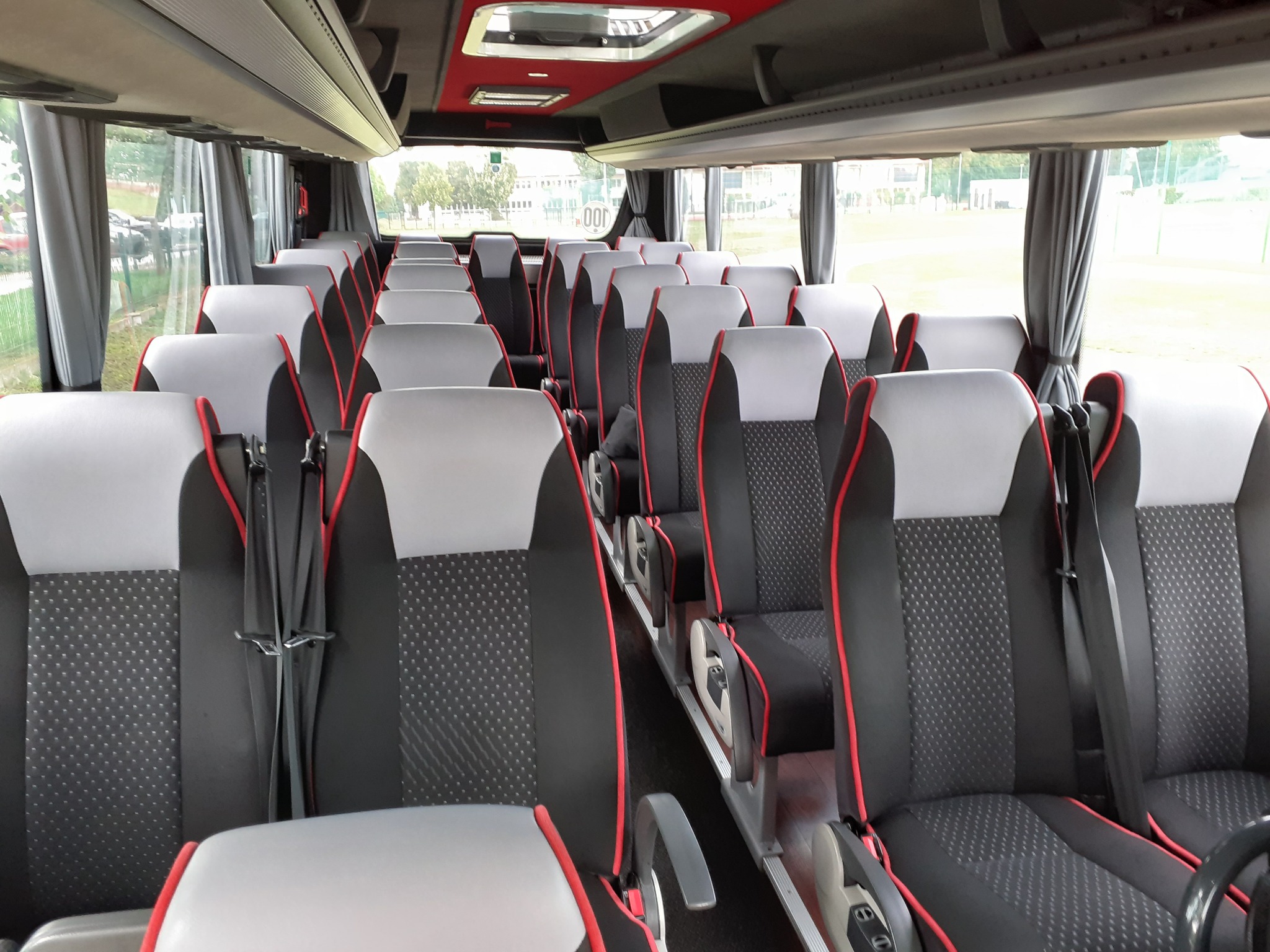 Wnętrze autobusu turystycznego z rzędami foteli w kolorze czarnym, szarym i czerwonym, widok z przodu pojazdu na puste siedzenia z pasami bezpieczeństwa.