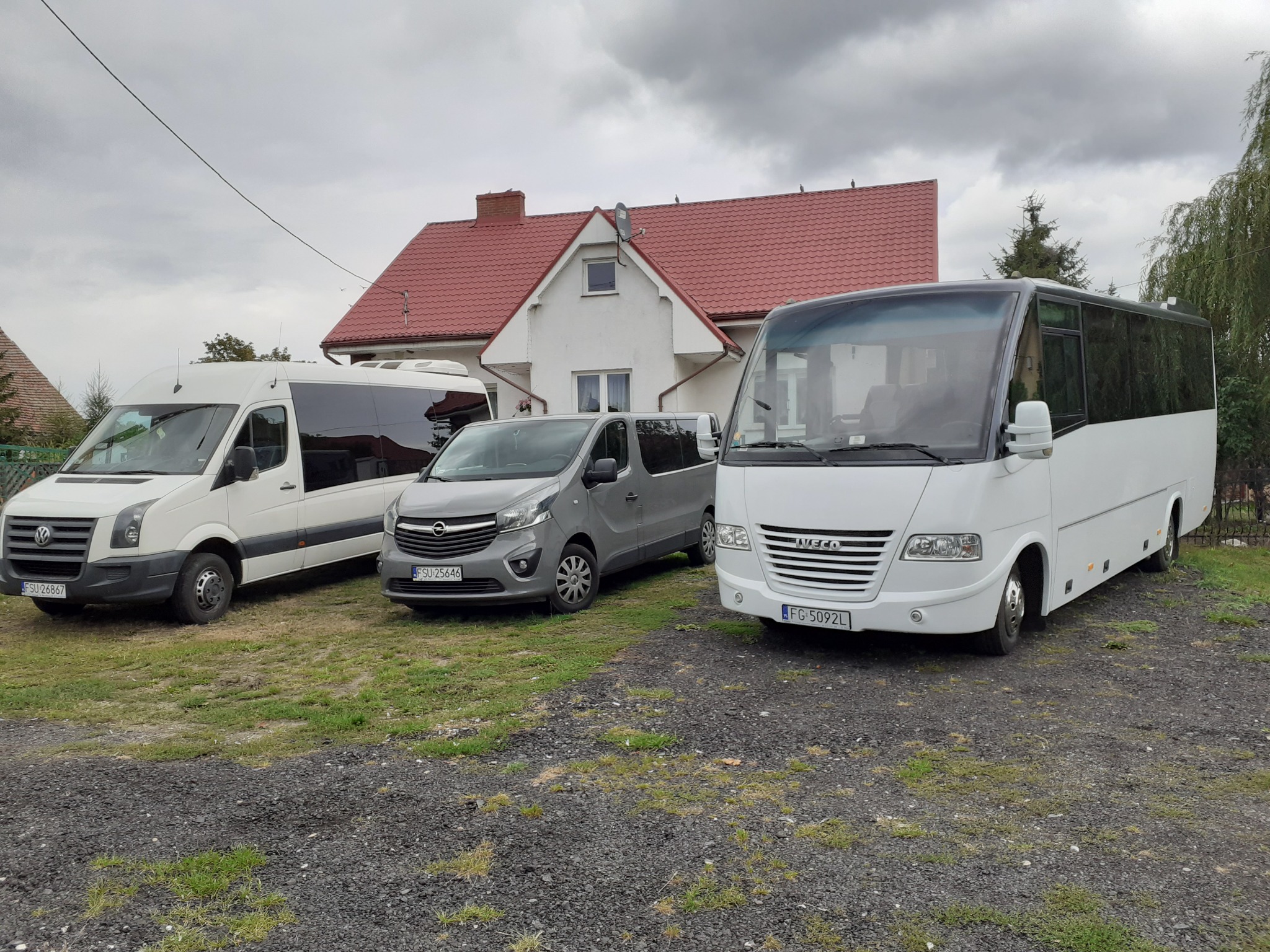 Flota trzech pojazdów: biały bus, szary van i biały minibus na żwirowym podłożu przed domem z czerwoną dachówką.