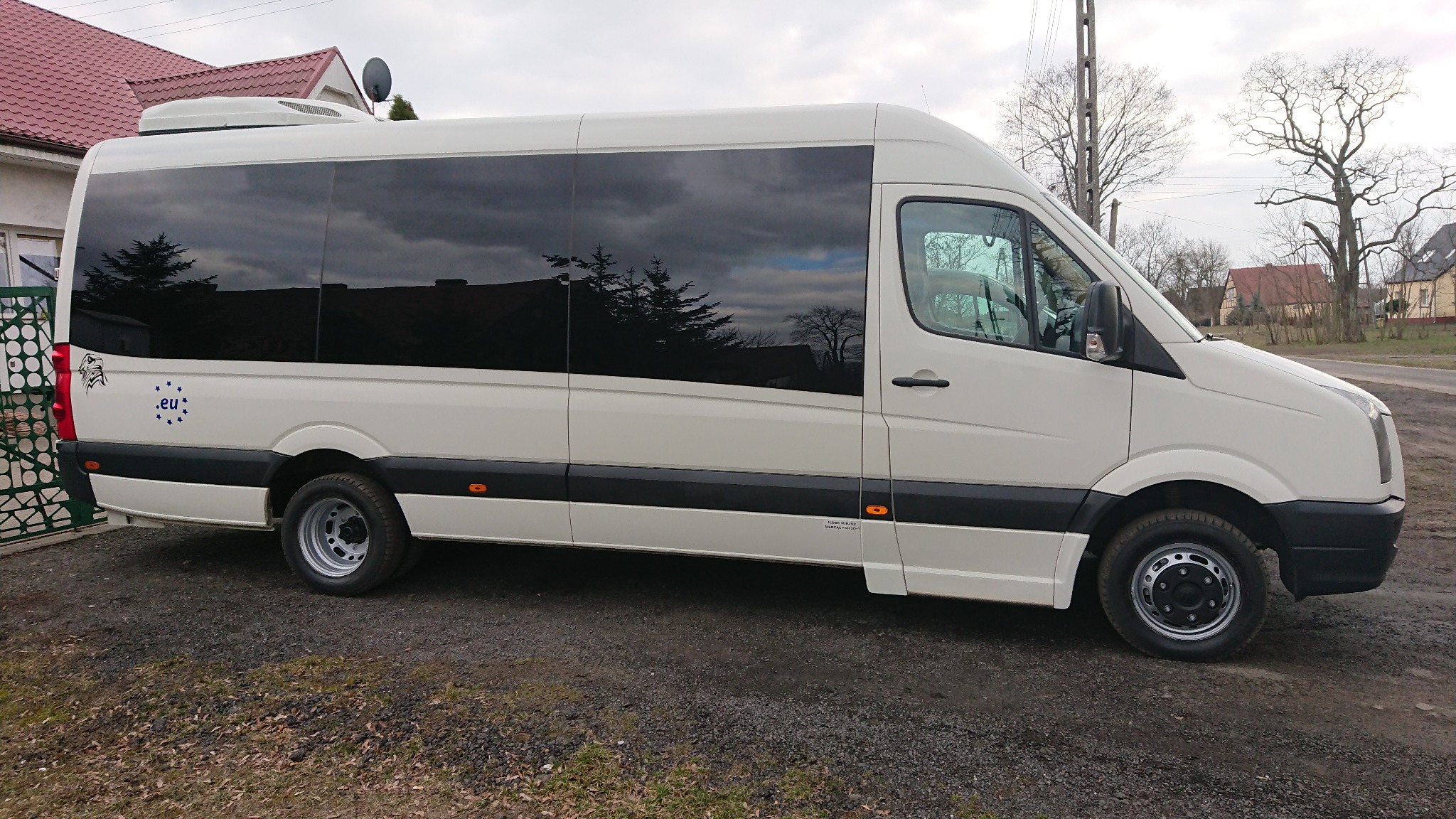 Biały bus z przyciemnianymi szybami i naklejką z gwiazdami UE, zaparkowany na żwirowym podłożu przed domem z czerwoną dachówką.