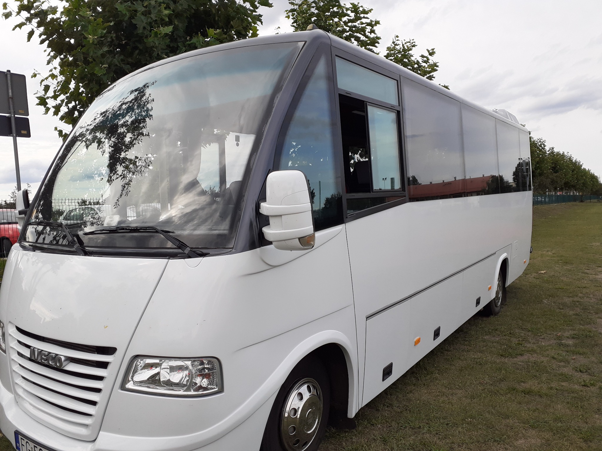 Biały, nowoczesny minibus Iveco na trawiastym terenie, widok z przodu i boku, z widocznym lusterkiem i przednią szybą odbijającą drzewa.