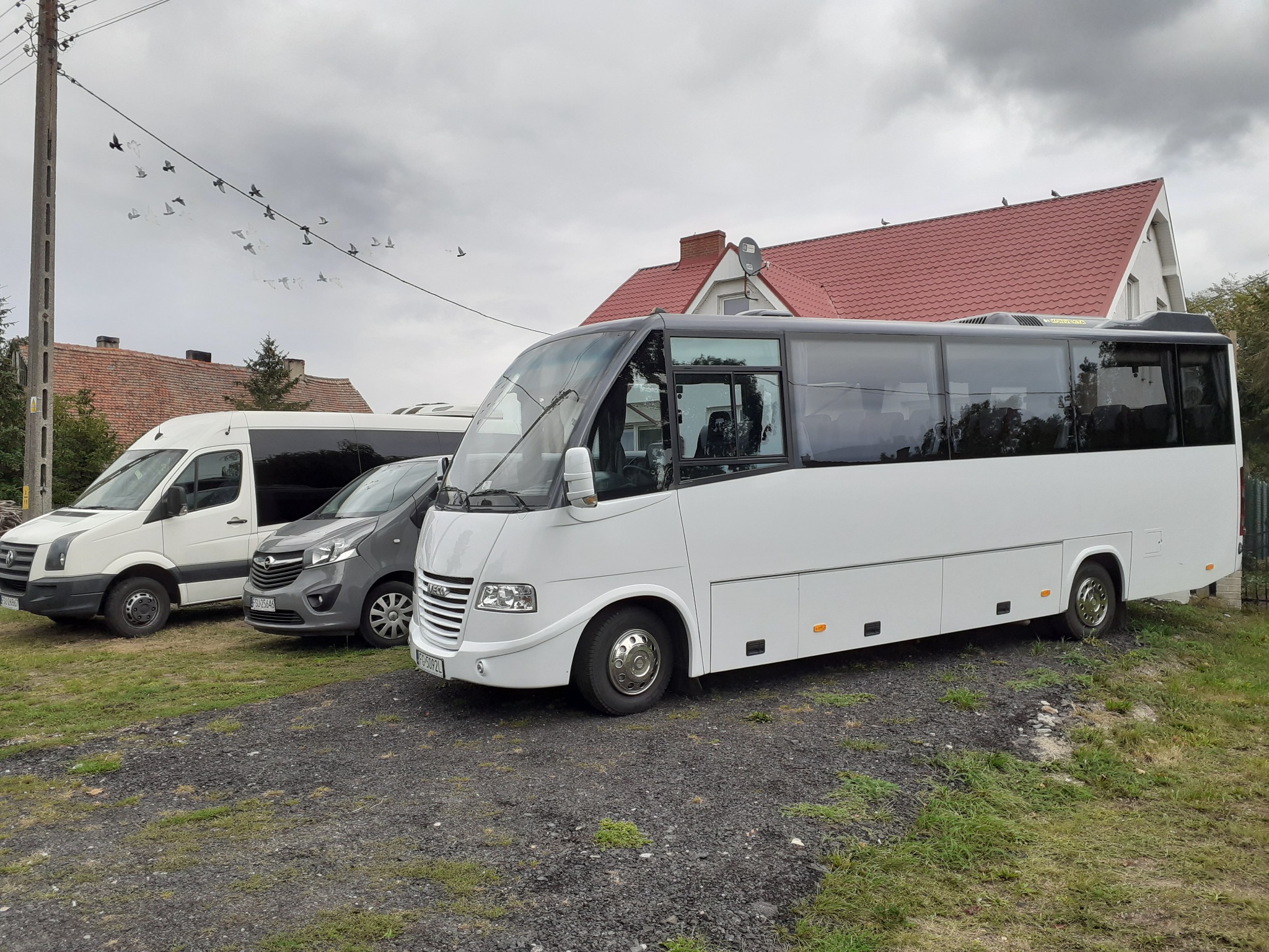 Flota pojazdów: biały bus, szary van i biały dostawczak zaparkowane na żwirowym podłożu przed budynkiem z czerwoną dachówką, stado ptaków na linii energetycznej.