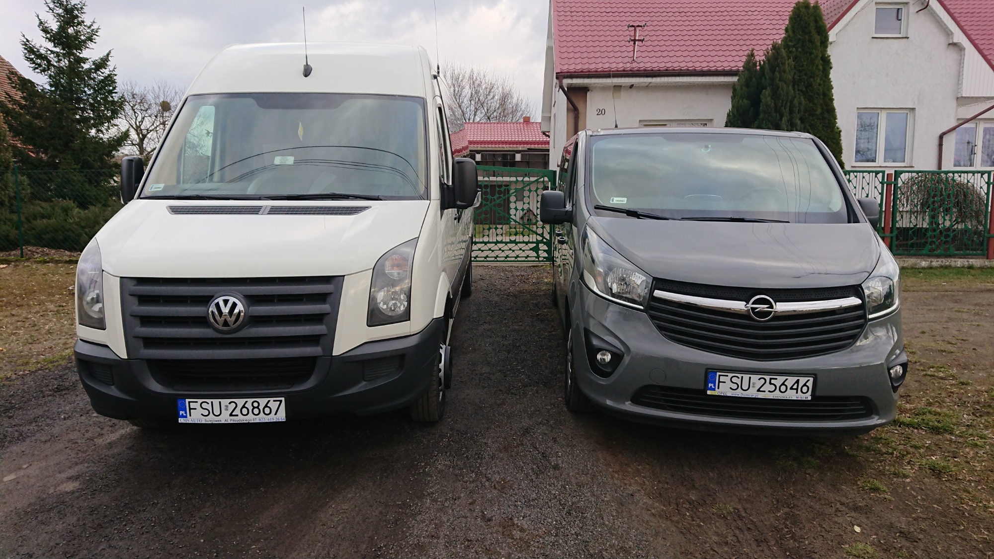 Dwa busy, biały Volkswagen Crafter i szary Opel Vivaro, zaparkowane obok siebie na żwirowym podjeździe przed domem z czerwonym dachem, widok z przodu.