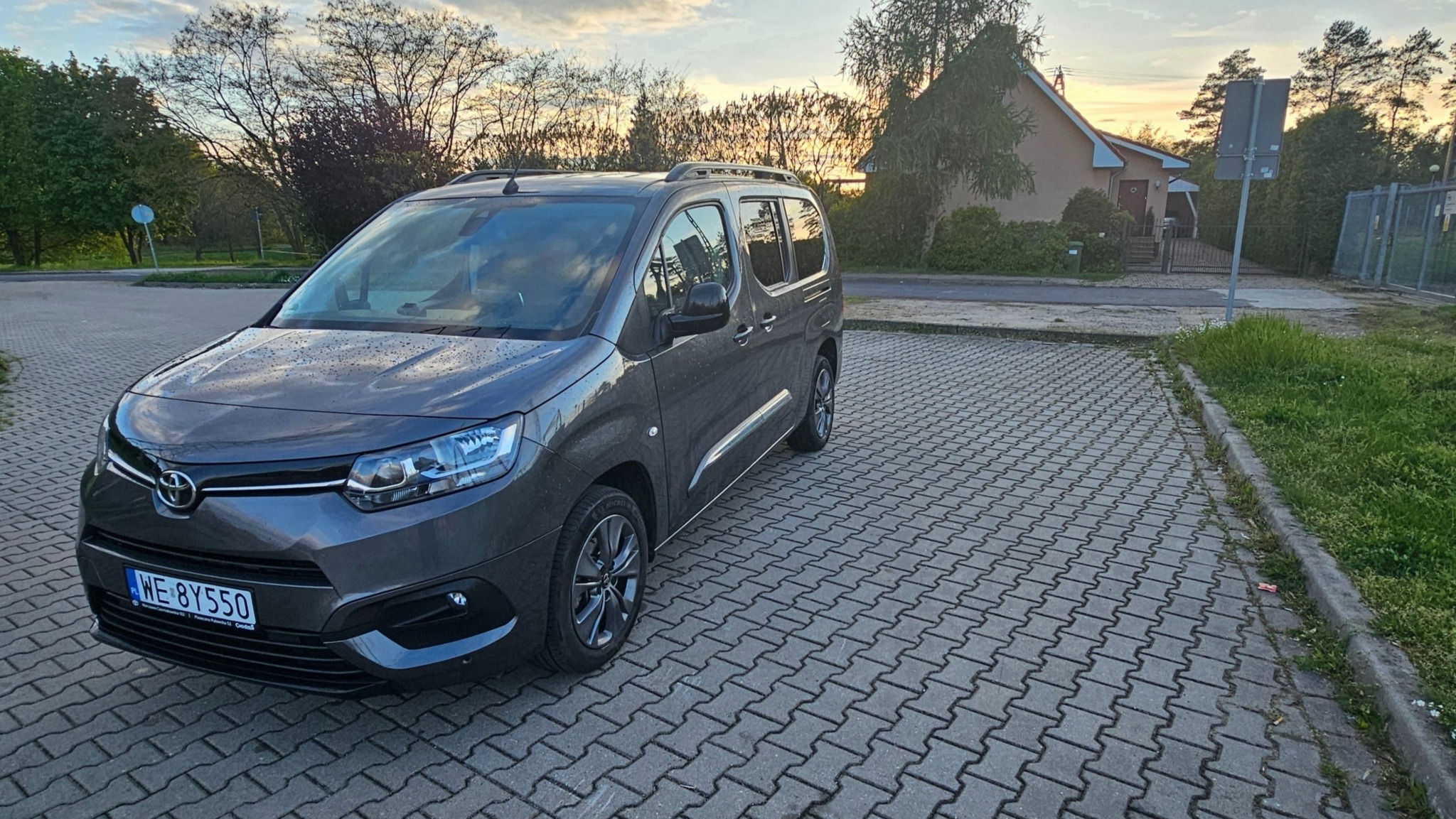 Szary samochód typu kombivan marki Toyota Proace City Verso z relingami dachowymi, zaparkowany na kostce brukowej, z widocznym numerem rejestracyjnym WE 8Y550, w tle dom i zachodzące słońce.