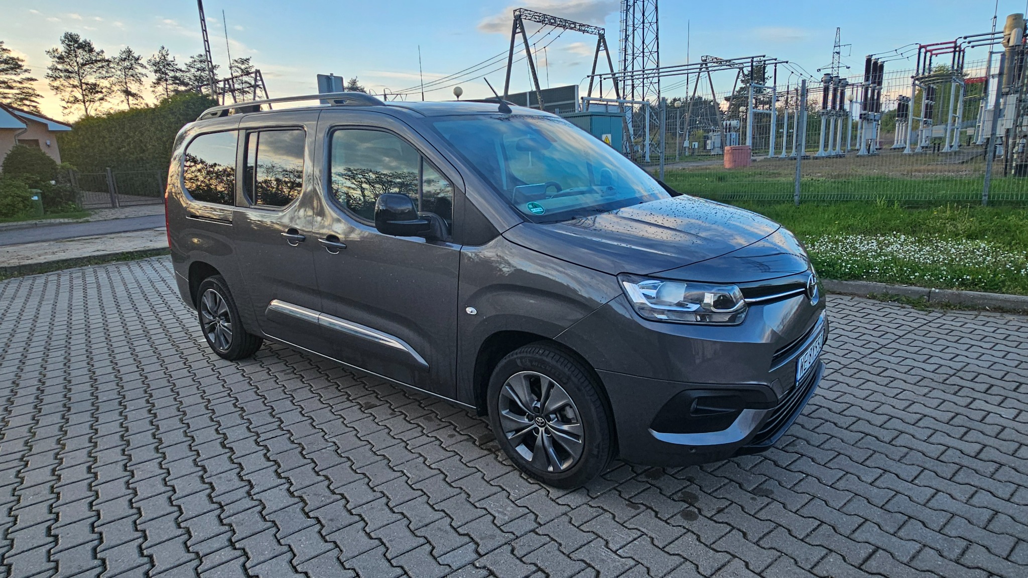 Ciemnoszary van z relingami dachowymi zaparkowany na kostce brukowej, w tle słupy wysokiego napięcia i zieleń.